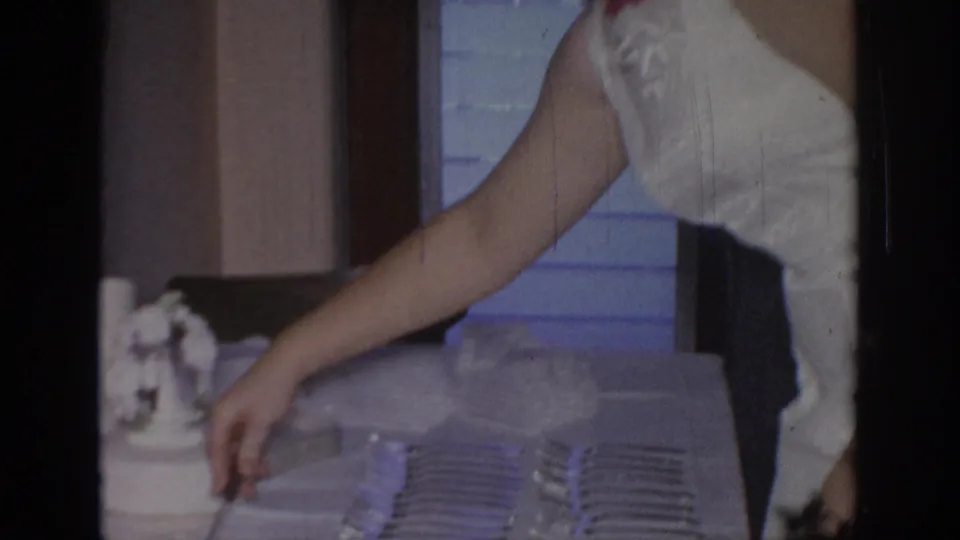 In a 1967 Oklahoma home-movie frame, a woman in a white sleeveless dress appears to arrange tableware on a serving surface.