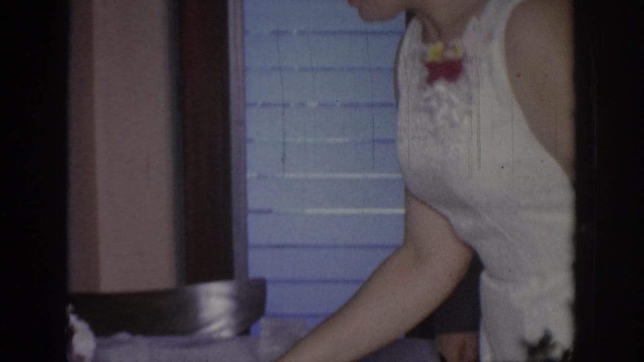 Woman in a white sleeveless dress bends near a table with window blinds behind her.