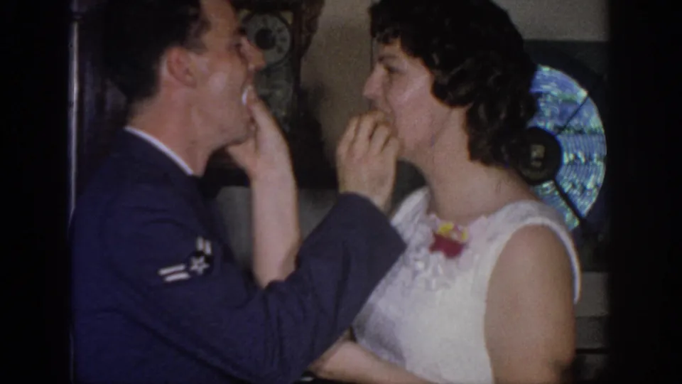 A man in a dark uniform and a woman in a white dress share cake in a close, playful 1967 Oklahoma home-movie moment.