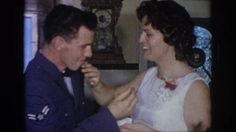 In 1967 Oklahoma home-movie footage, a uniformed man and bride smile while sharing cake in a private interior.
