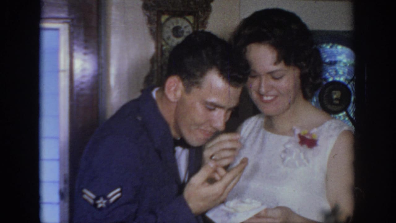 The bride smiles beside a uniformed man examining cake, with a clock and decorative fan behind them.