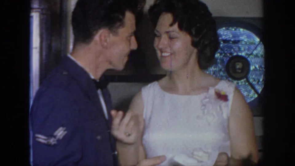 The couple faces each other smiling, hands clasped, in a private 1960s wedding reception interior.