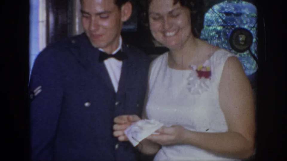 A smiling Air Force serviceman stands beside the woman in a white dress as they hold or examine a paper in 1967 Oklahoma footage.