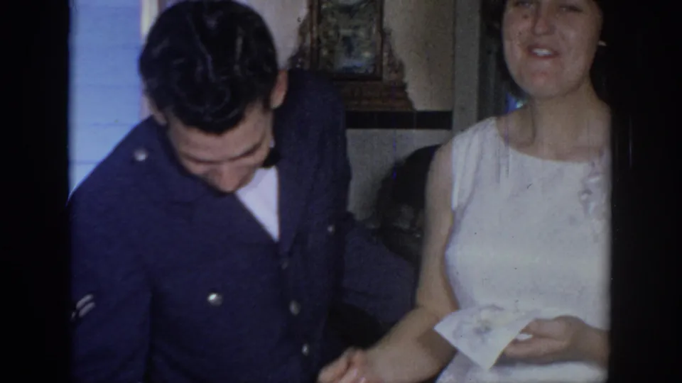 The serviceman looks downward as the woman in a white dress holds the small paper, with interior wedding surroundings partly visible.