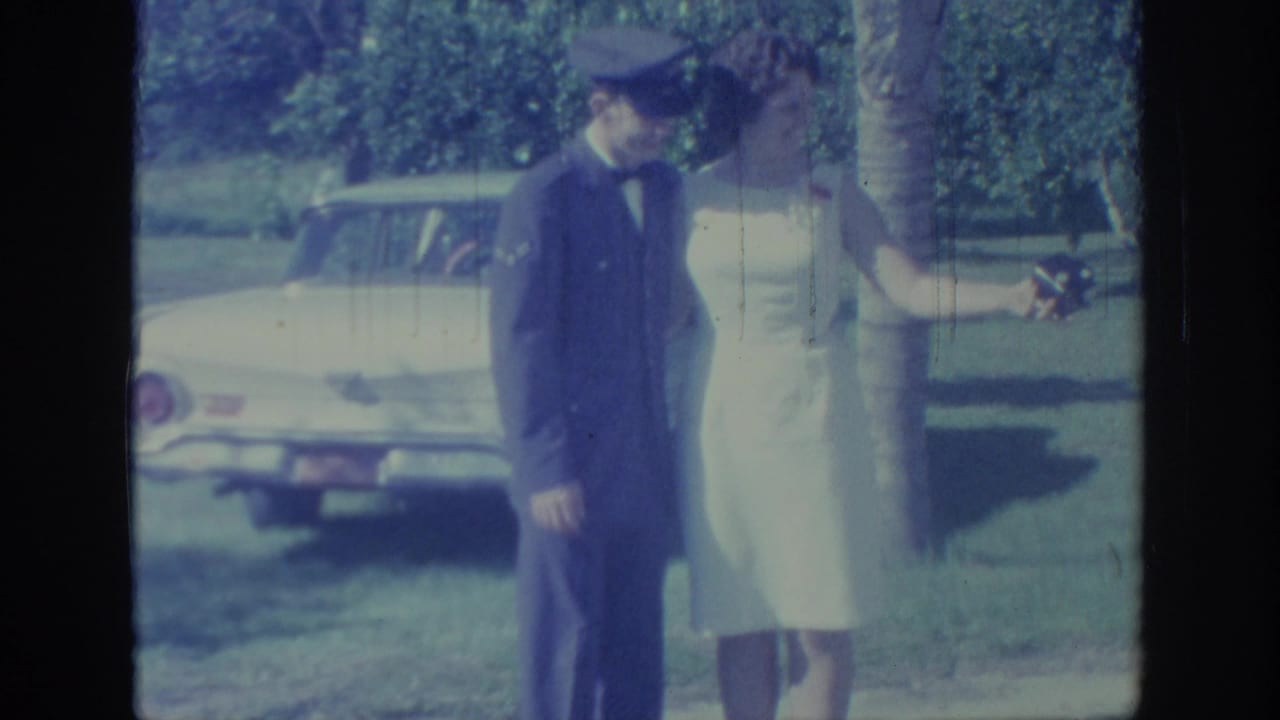 In 1967 Oklahoma home-movie footage, a man in a dark uniform and a woman in a white dress stand near a light-colored car on a sunny lawn.