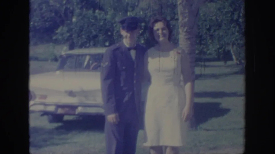 The couple faces the camera in 1967 Oklahoma footage, standing on grass before a light-colored 1960s car and trees.