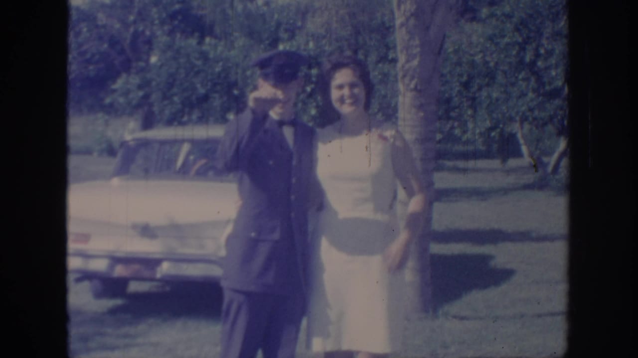 1967 Oklahoma 8mm frame showing a uniformed man waving beside a woman in a light dress, with a parked car, grass, and trees behind them.