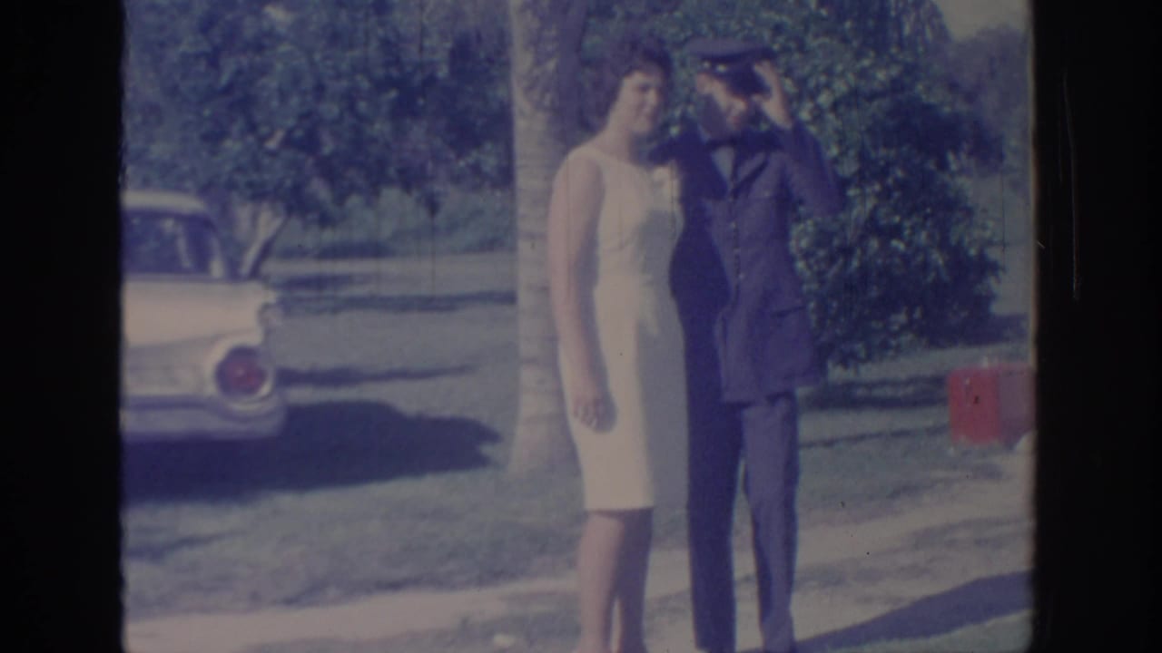 Woman in a light dress stands beside a uniformed man saluting outdoors near a parked car.