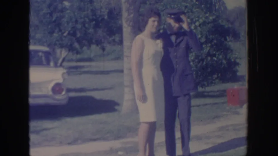 1967 Oklahoma 8mm frame of a woman in a light sleeveless dress beside a uniformed man raising a salute near a parked 1960s car.