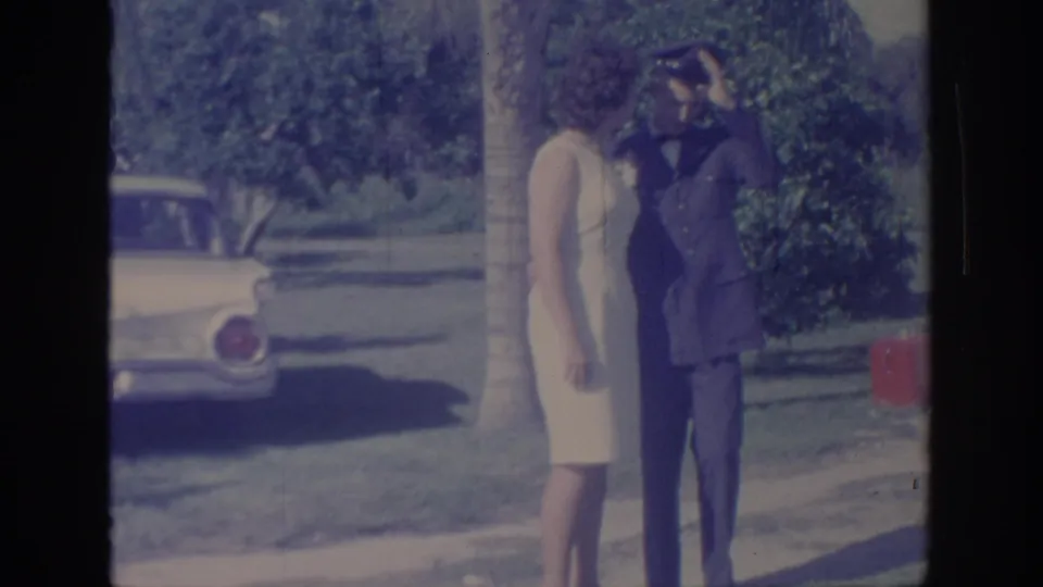 1967 Oklahoma home-movie frame showing a uniformed man saluting next to a woman in a light dress on a lawn near a parked car.
