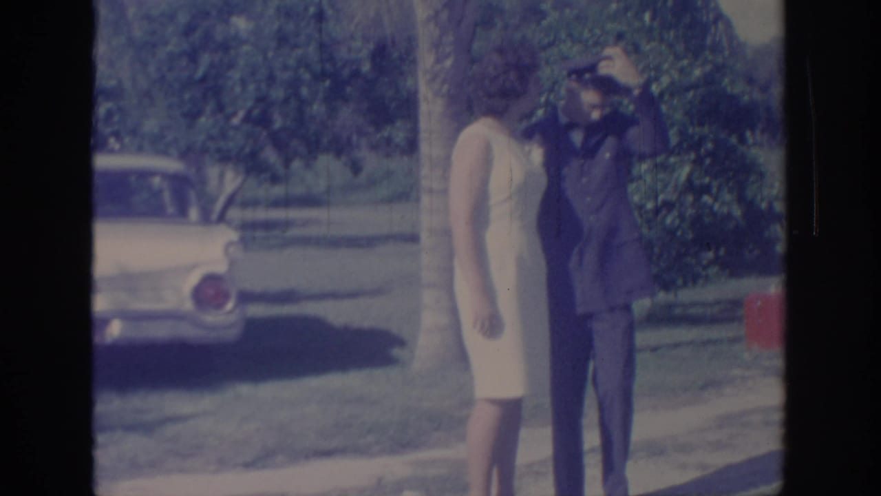 In 1967 Oklahoma home-movie footage, a woman in a cream dress adjusts a man’s military cap near a classic car in a sunny park setting.