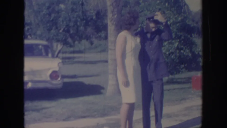 In 1967 Oklahoma home-movie footage, a woman in a cream dress adjusts a man’s military cap near a classic car in a sunny park setting.