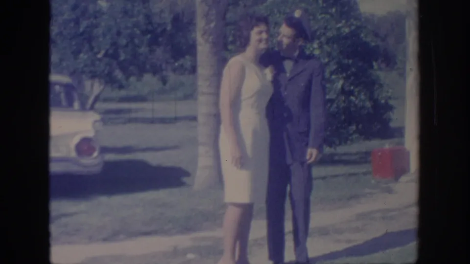 The couple poses close together in a sunlit Oklahoma park, with the man in uniform and a mid-century car behind them.