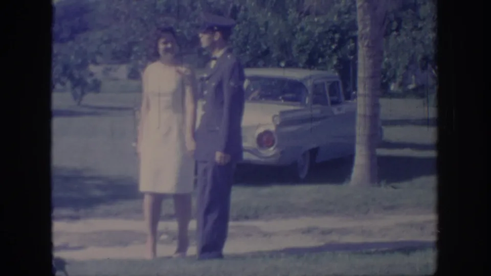 A woman in a light dress and a man in military uniform stand facing each other near a classic 1960s car in Oklahoma home-movie footage.