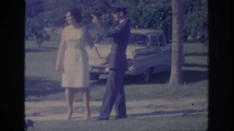 The couple walks hand-in-hand past the parked classic car, with trees and lawn behind them in the 1967 Oklahoma home-movie scene.