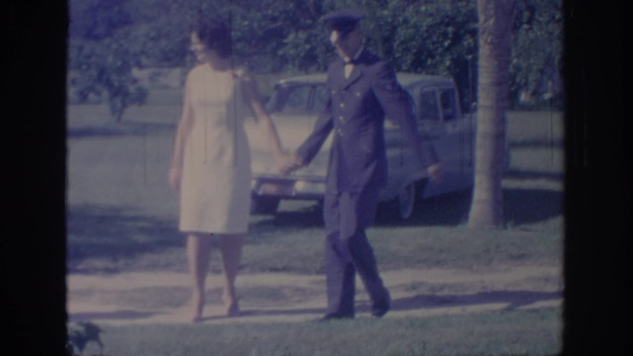 In 1967 Oklahoma home-movie footage, a woman in a white dress and a man in a dark uniform hold hands outdoors near a parked car.