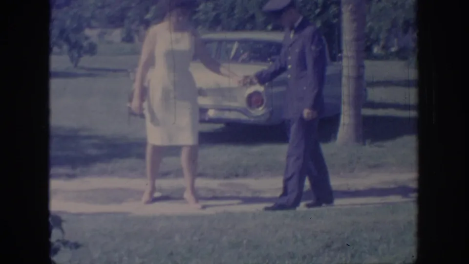 A man in a dark uniform faces a woman in a white sleeveless dress during a casual outdoor home-movie moment in Oklahoma.