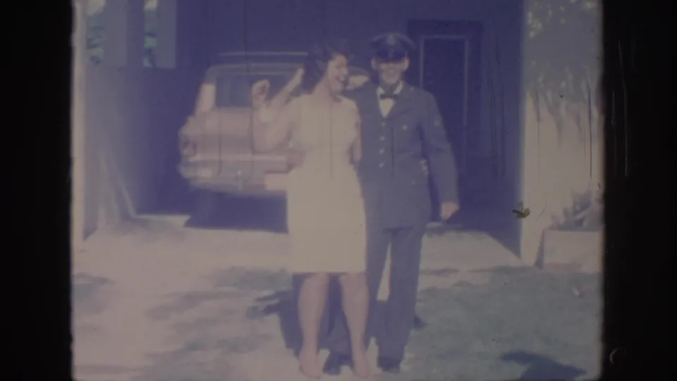 The couple poses close together near a driveway or house area, with the woman in a white dress beside the uniformed man.
