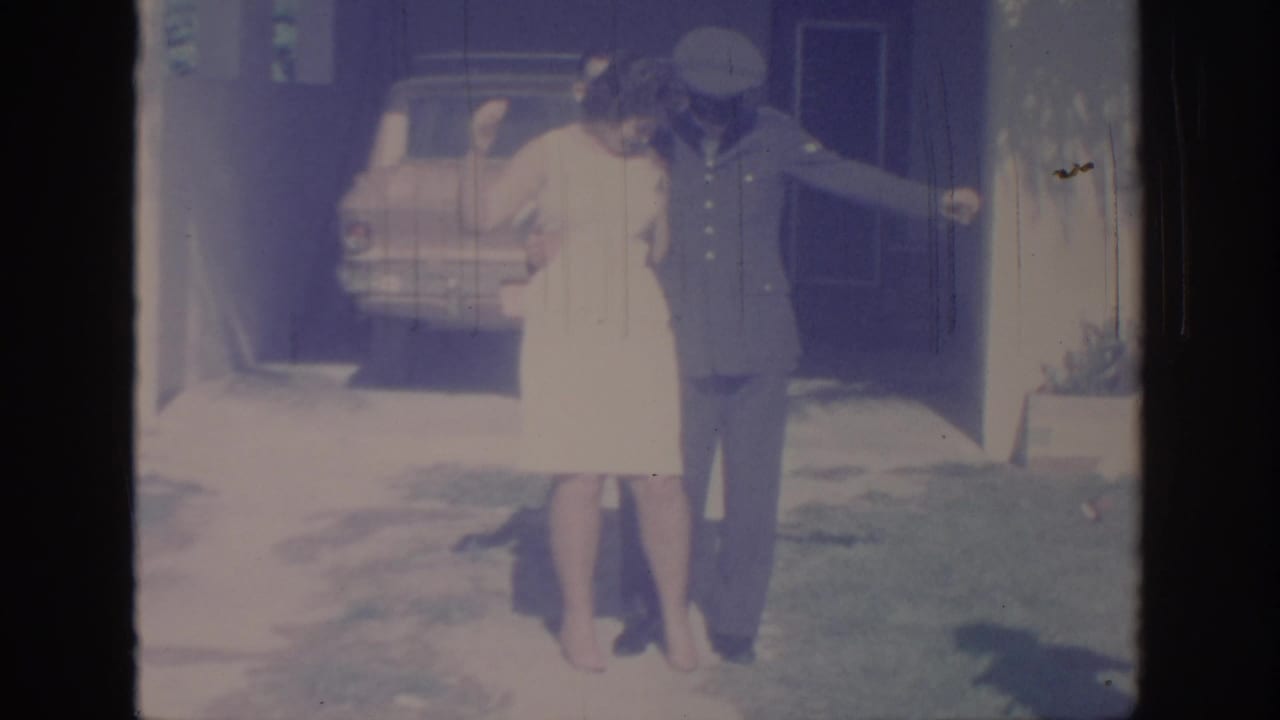 1967 Oklahoma home-movie frame showing a woman posing with a uniformed man near a parked car and house entry.
