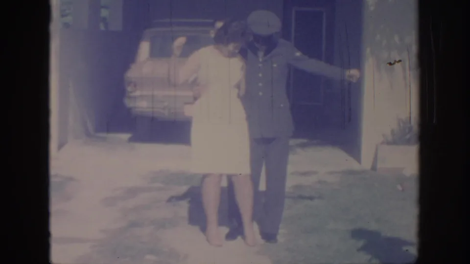 1967 Oklahoma home-movie frame showing a woman posing with a uniformed man near a parked car and house entry.