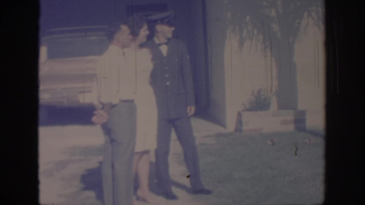 1967 Oklahoma home-movie frame of a uniformed soldier standing with two adults on a lawn beside a house and parked car.
