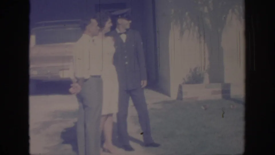 1967 Oklahoma home-movie frame of a uniformed soldier standing with two adults on a lawn beside a house and parked car.