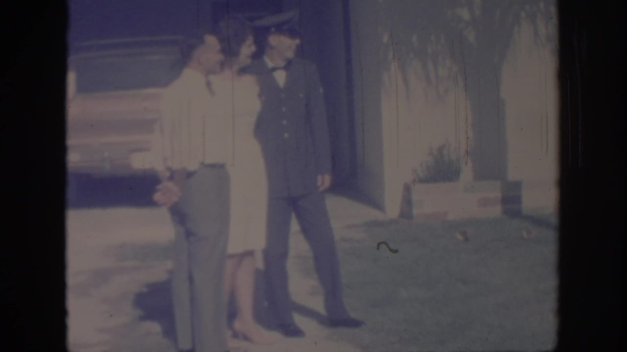 A uniformed soldier and two adults stand close together on a sunny lawn outside a house in 1967 Oklahoma footage.