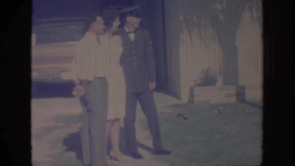 A uniformed soldier and two adults stand close together on a sunny lawn outside a house in 1967 Oklahoma footage.