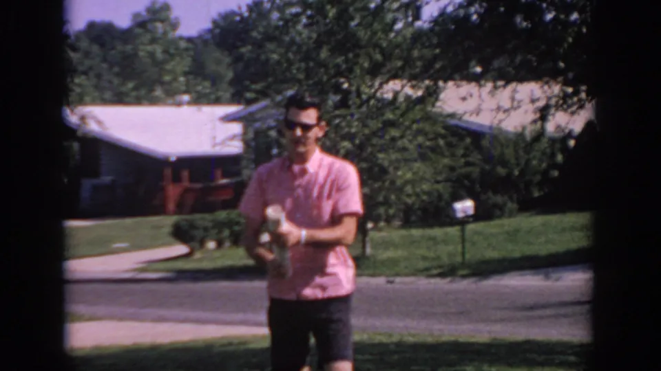 In 1967 Oklahoma home-movie footage, a young man in a pink short-sleeve shirt holds a rolled newspaper on a sunny suburban lawn.