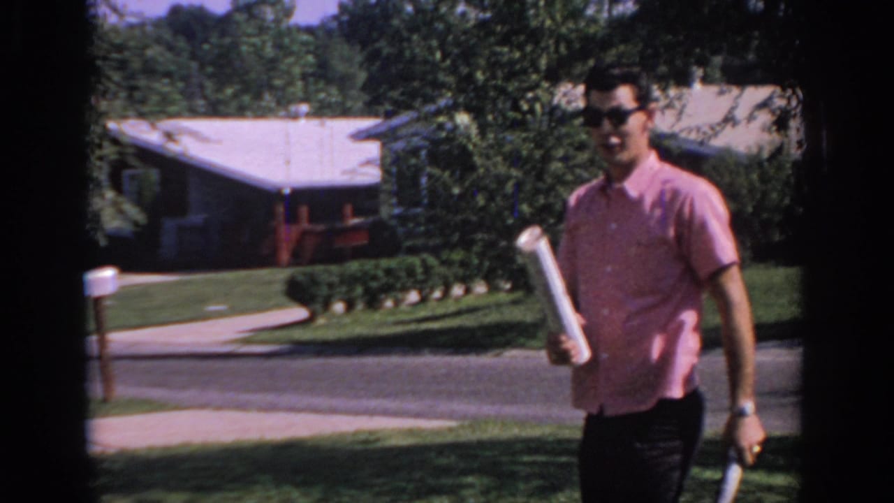 Young man in sunglasses walks near the camera holding a rolled newspaper beside a front lawn.