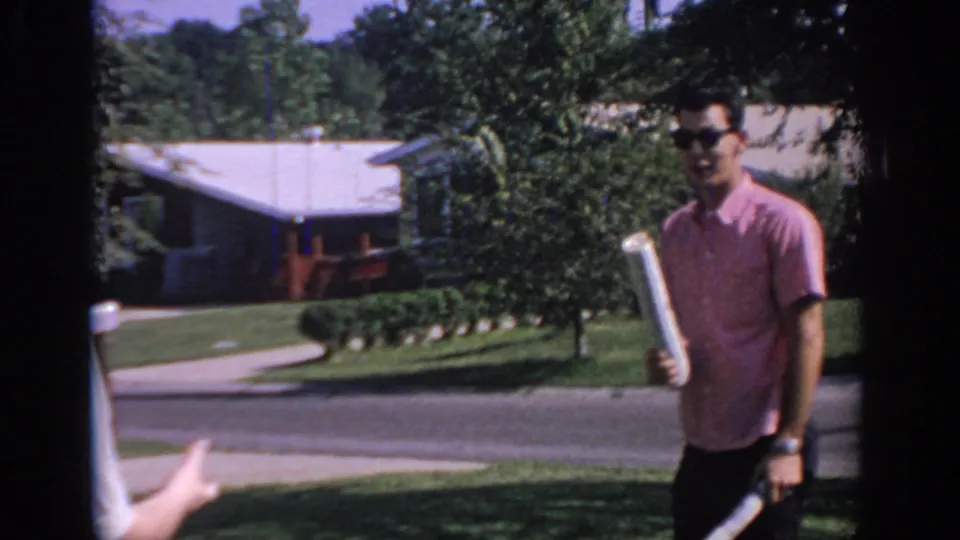 The young man stands with the newspaper while another person's hand reaches into frame, suggesting a playful exchange on the lawn.