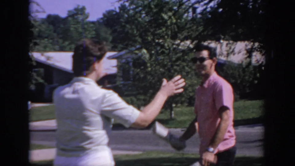 A woman in a light dress reaches toward the young man with the newspaper during a playful 1967 Oklahoma home-movie moment.