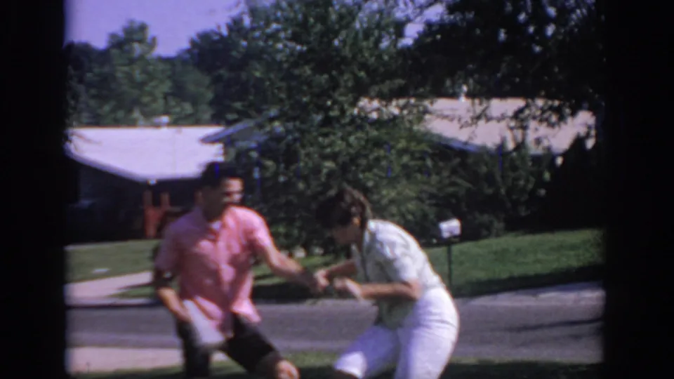 The man in pink and woman in white bend and laugh while pulling the newspaper on a quiet Oklahoma street in 1967 footage.