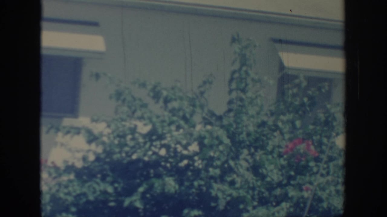 1967 Oklahoma 8mm frame showing a green yard bush before a pale house wall, rectangular window, and white awning.
