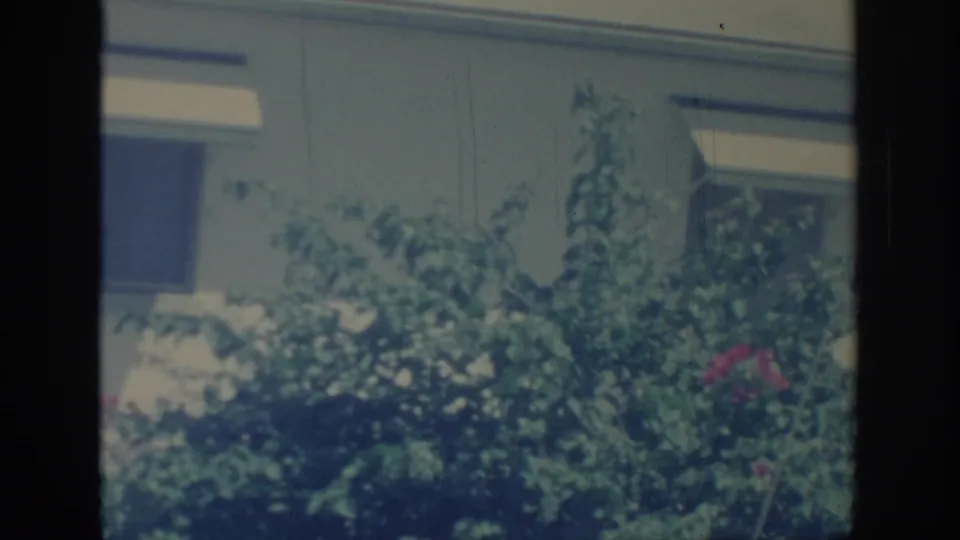 1967 Oklahoma 8mm frame showing a green yard bush before a pale house wall, rectangular window, and white awning.