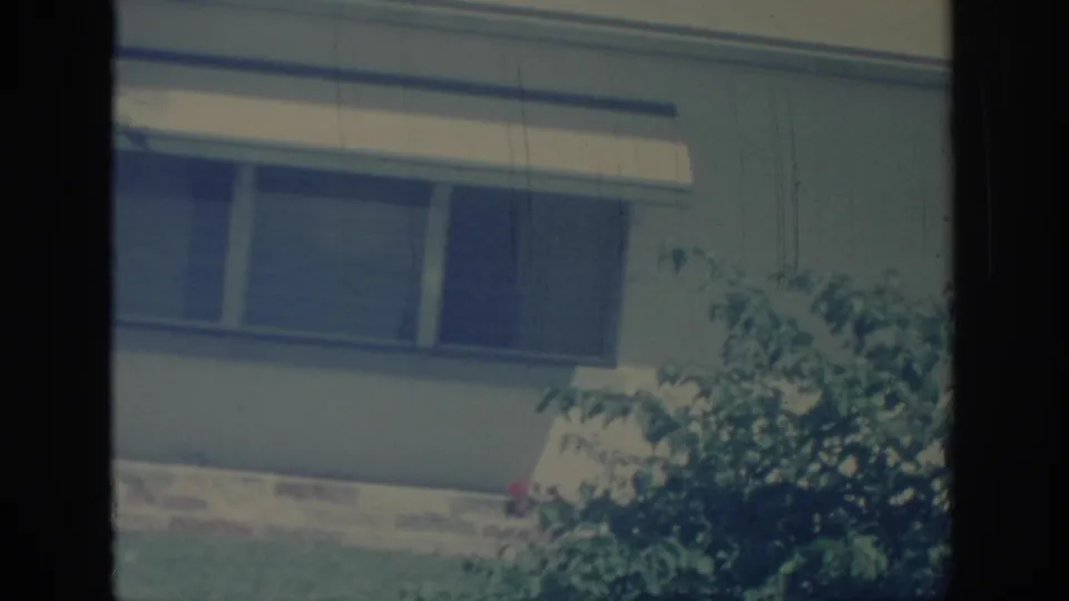 1967 Oklahoma 8mm frame showing a house window, awning, lawn, brick base, and shrub with small red flowers.