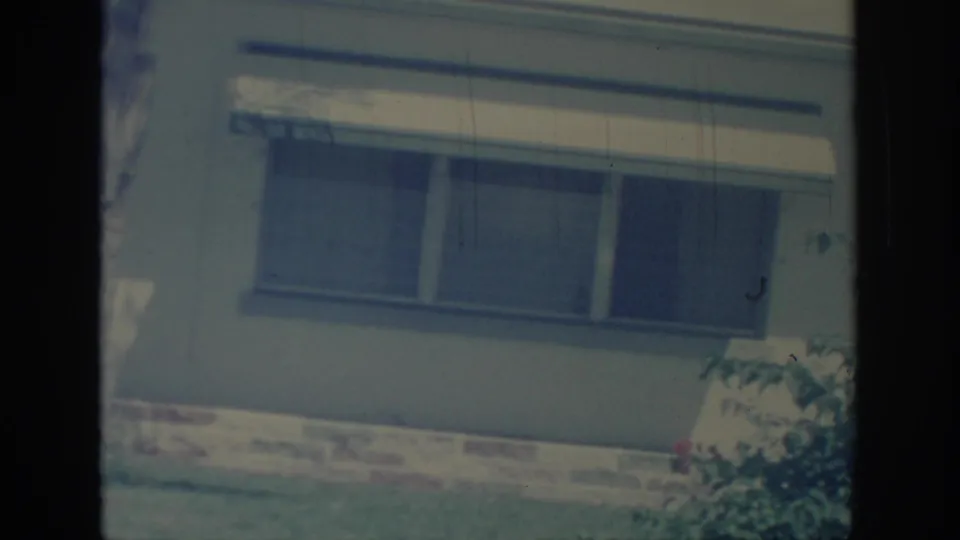 1967 Oklahoma home-movie frame with a pale house wall, shaded window, white awning, brick foundation, lawn, and shrub edge.