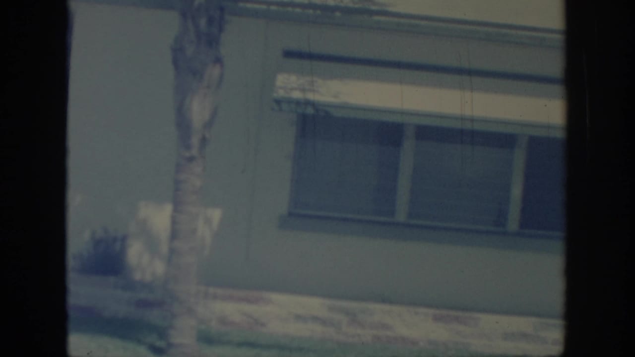 1967 Oklahoma 8mm frame showing a single-story house exterior with a shaded window, palm tree trunk, lawn, and garage edge.