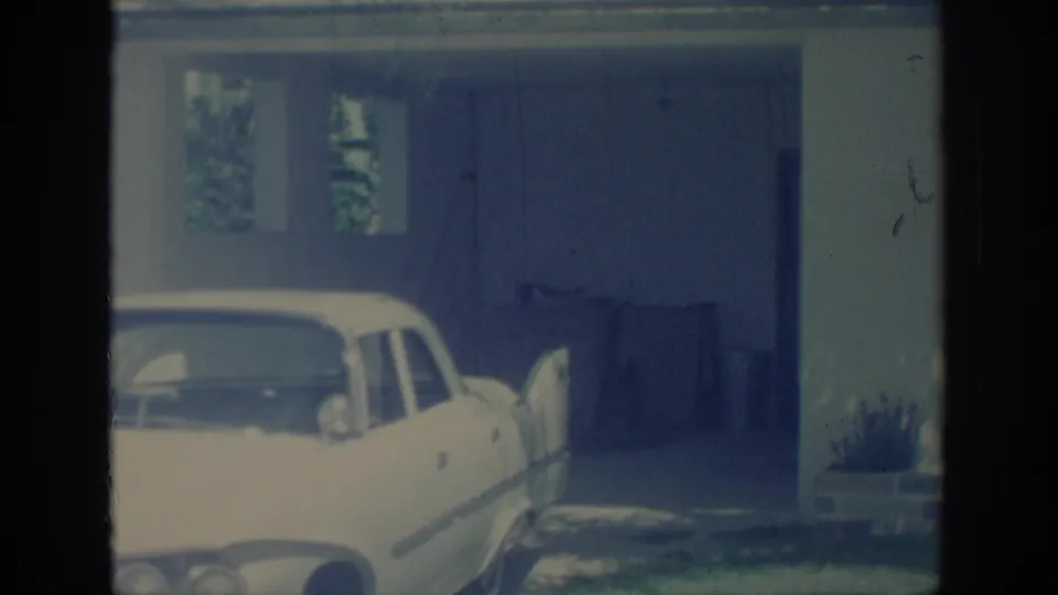 1967 Oklahoma frame with a white car parked in front of a covered garage, bins, house wall, and small windows.