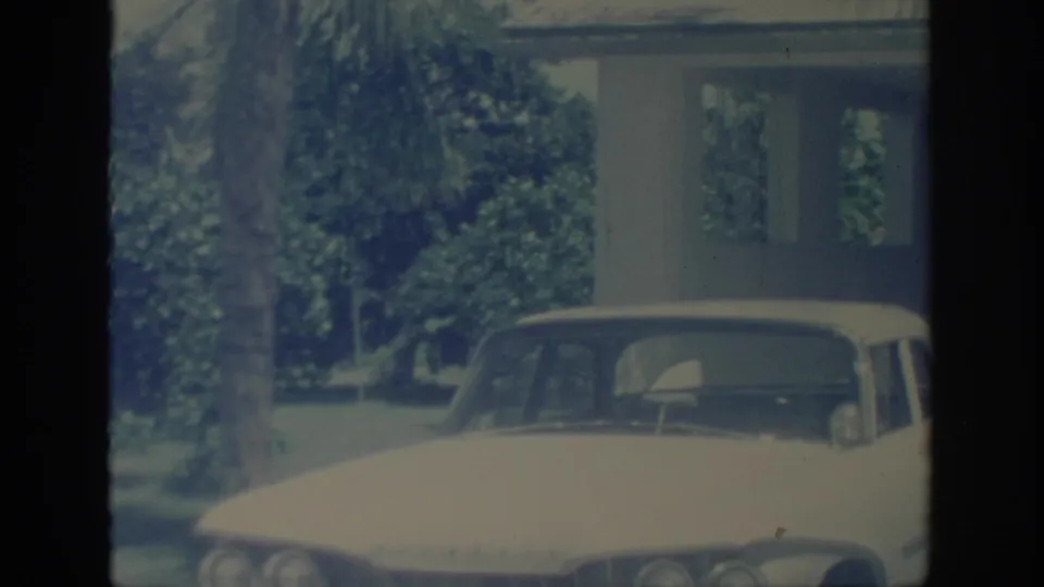 1967 Oklahoma 8mm frame with the front of a white car, a residential carport, lawn, trees, and a tall palm-like trunk.