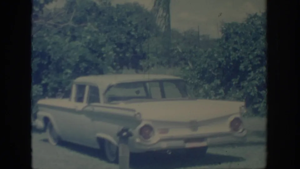 1967 Oklahoma 8mm frame showing a vintage sedan from the rear quarter with chrome trim and dense green foliage behind it.