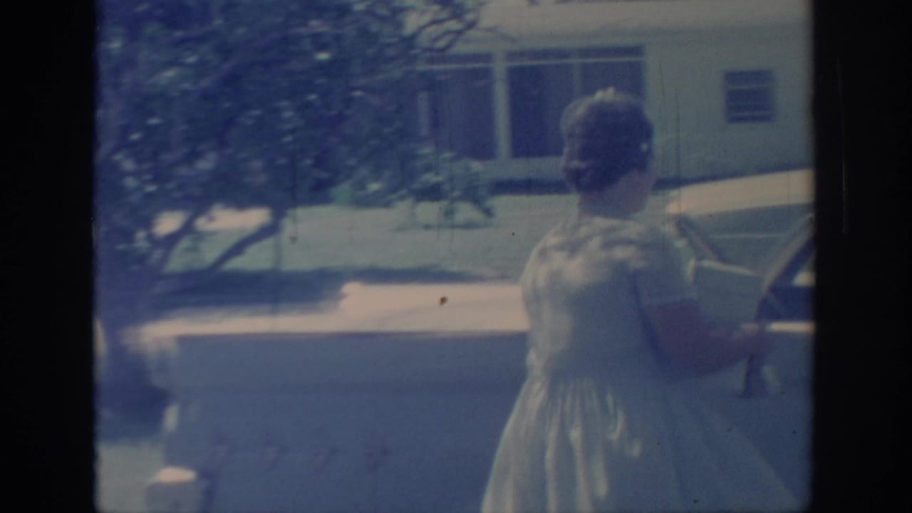 The woman continues toward a parked finned car on a sunlit suburban street in the 1967 Oklahoma footage.
