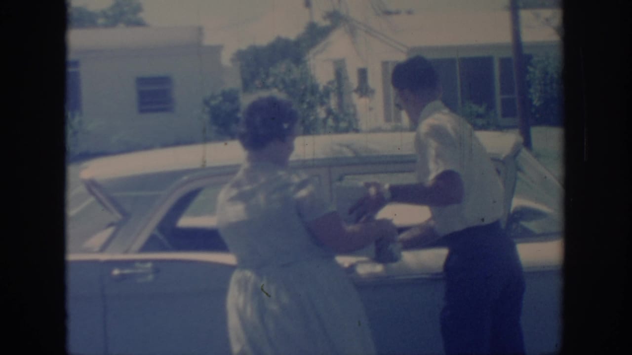 In 1967 Oklahoma home-movie footage, a woman in a pale dress and a man in dark trousers stand at the rear side of a parked sedan.