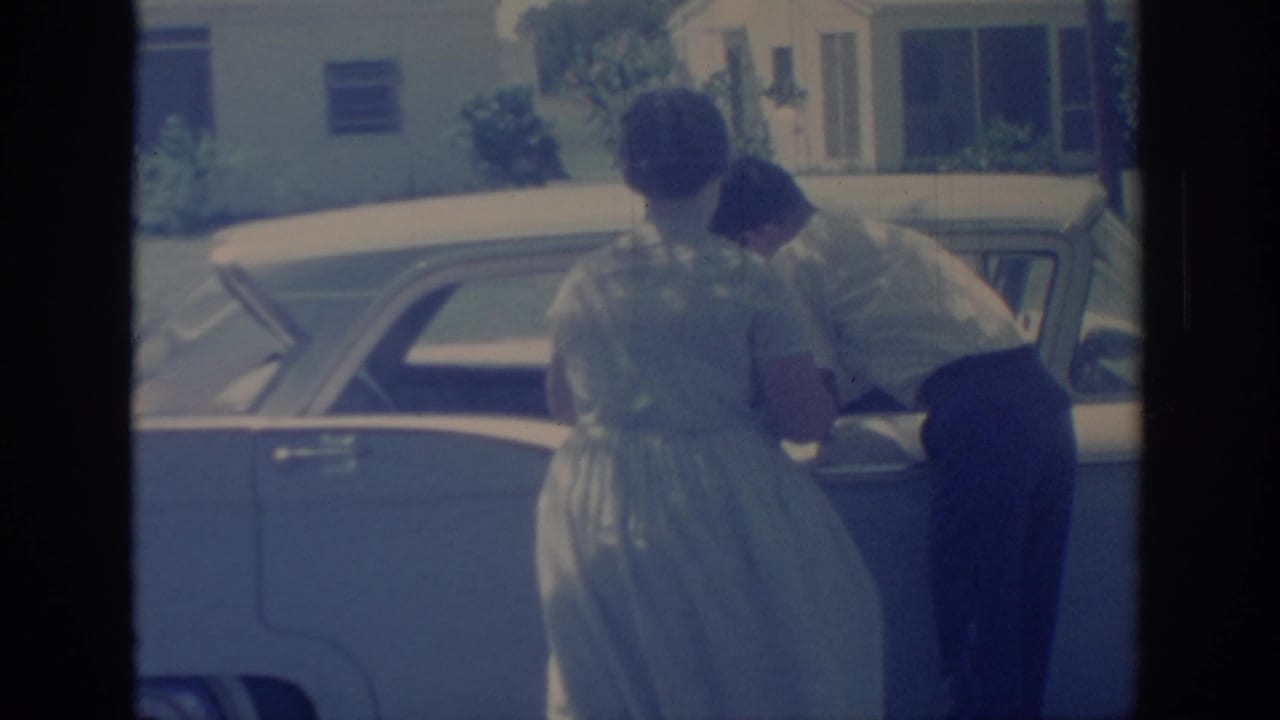 A woman in a light dress watches as the man bends toward the rear side of the mid-century sedan in this faded 8mm frame.