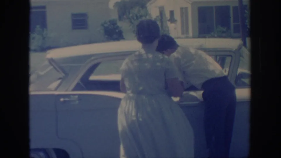 A woman in a light dress watches as the man bends toward the rear side of the mid-century sedan in this faded 8mm frame.