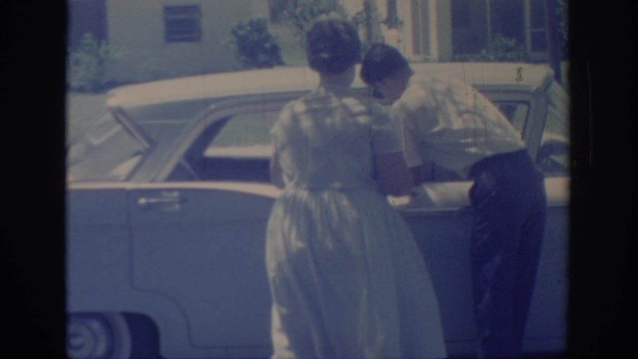 The man appears to retrieve or place something at the sedan's rear side as the woman stands nearby on a sunny Oklahoma street.