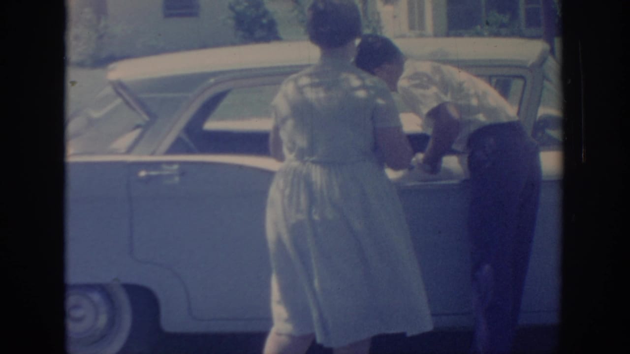 In 1967 Oklahoma home-movie footage, a woman in a pale dress and a man in dark trousers stand beside a light-colored sedan.