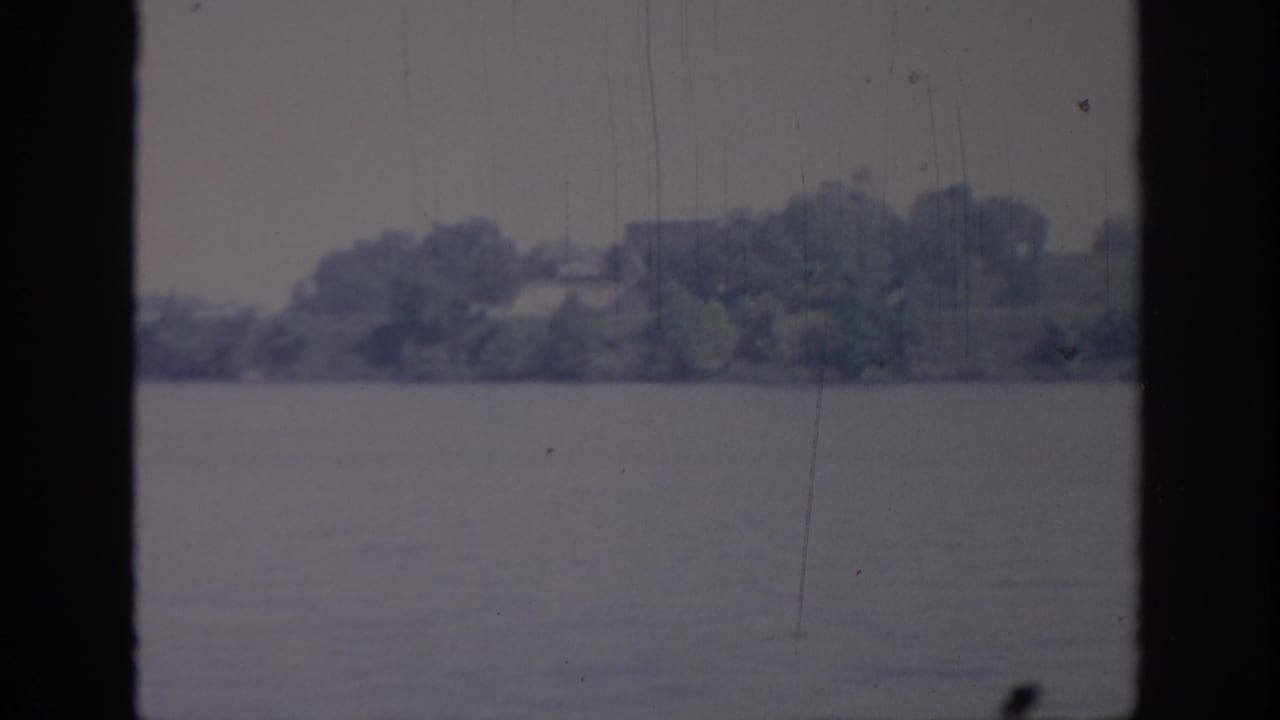 1967 Oklahoma 8mm frame showing calm lake water, a tree-lined far shore, and dark vertical window framing.
