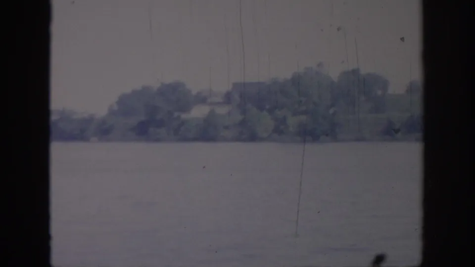 1967 Oklahoma 8mm frame showing calm lake water, a tree-lined far shore, and dark vertical window framing.