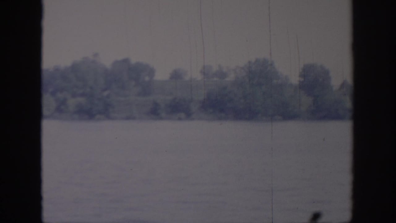 1967 Oklahoma home-movie frame with a soft tree-covered lakeshore, still water, and aged 8mm film texture.